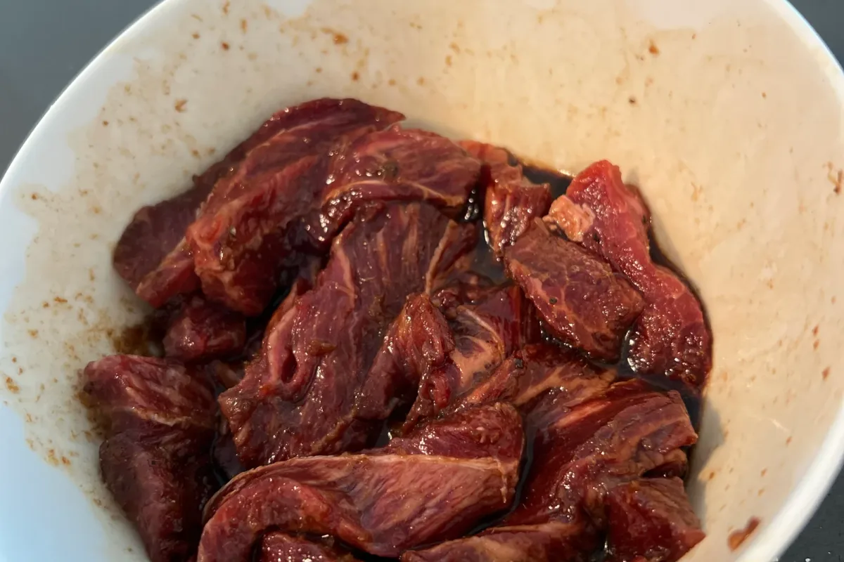 Sliced beef strips marinating in a dark sha cha and soy sauce mixture in a white bowl.