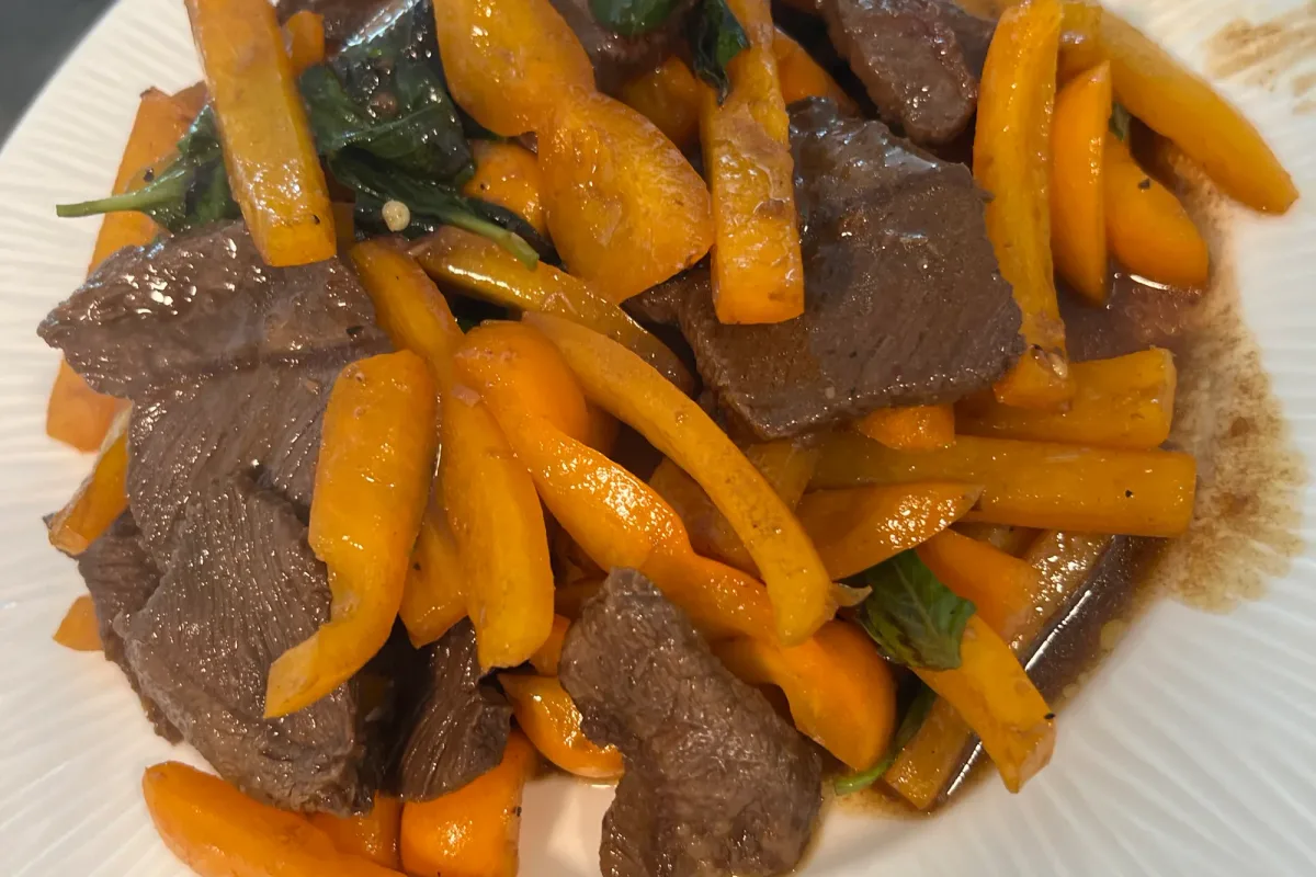 A plate of stir-fried flat iron steak and orange bell peppers glazed in sha cha sauce, topped with wilted Thai basil, on a ribbed white plate.